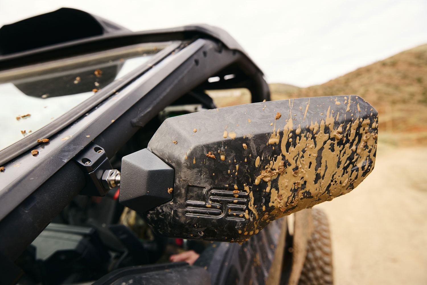 Mud-covered forged carbon fiber UTV side mirror mounted on roll cage, showing trail-tested durability in off-road conditions